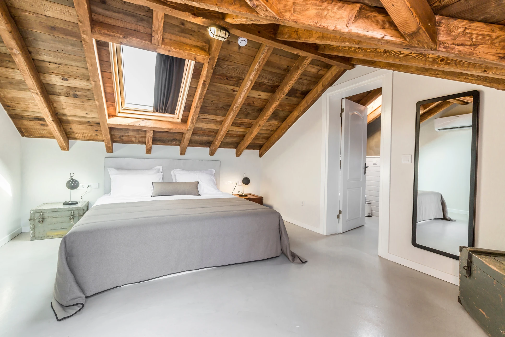 Chambre moderne sous combles avec poutres apparentes en bois naturel, lit double avec linge gris et blanc, sol béton ciré gris clair