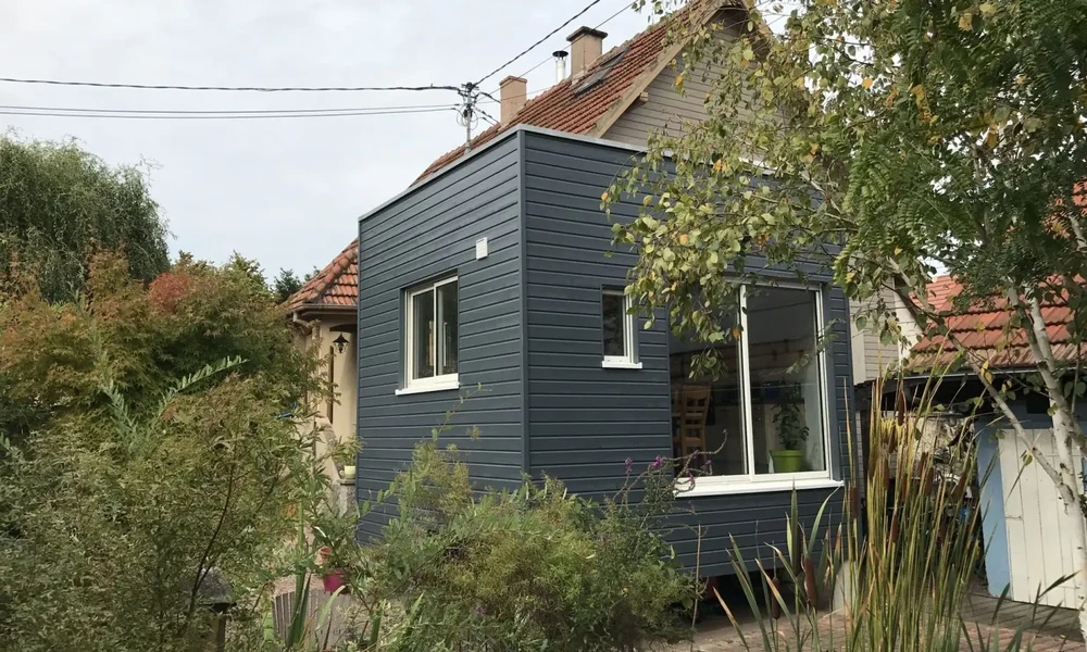 Scène extérieure résidentielle montrant une extension moderne à bardage horizontal bleu-gris avec fenêtres blanches, accolée à une maison traditionnelle à toiture tuiles rouges